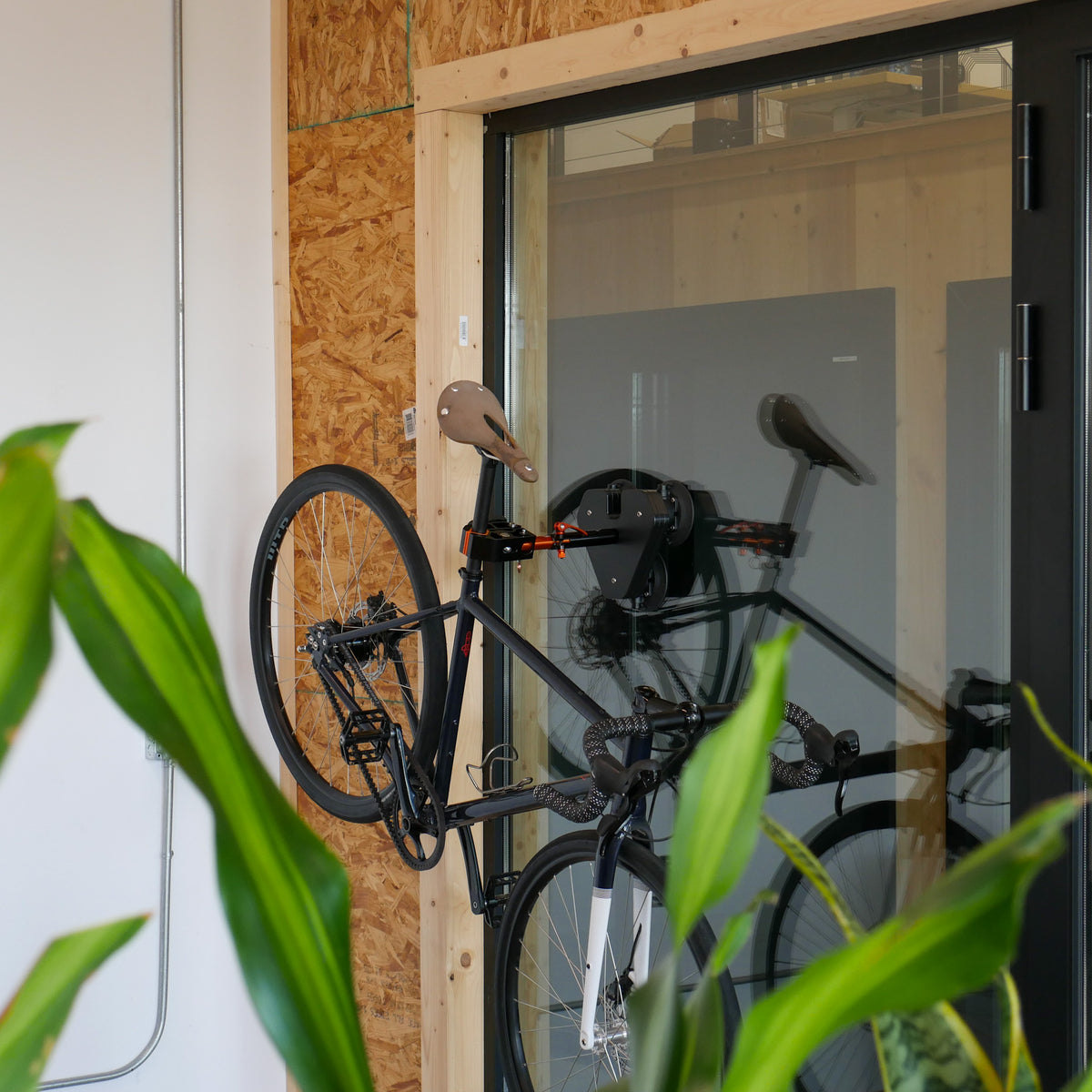 A Connect Air bike stand mounted on a glass wall inside a modern office, securely holding a Priority Apollo Gravel Bike by the seat post. The environment features wooden and glass architectural elements, showcasing the stand's versatility and compatibility with stylish interior decor