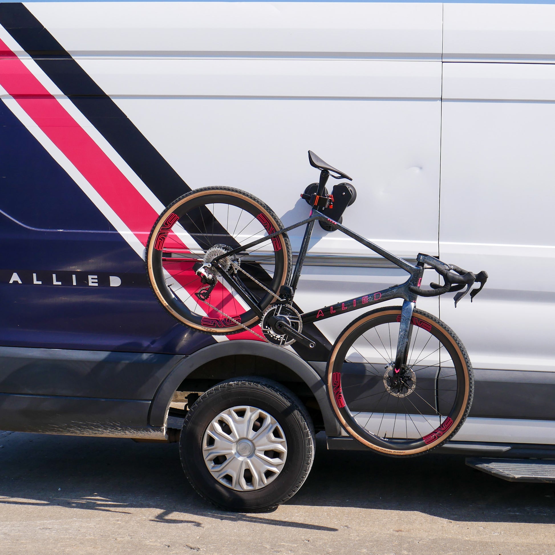 A Connect Air bike stand mounted on the side of an Allied branded vehicle securely holds a Allied Echo road gravel bike. The bike, featuring a striking pink and black color scheme, is displayed prominently against the vehicle’s reflective silver and blue striped design, demonstrating the stand’s versatility and strong visual appeal