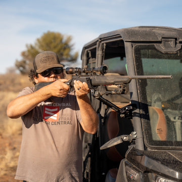 Hunter steadying a bolt-action rifle on a Connect Aim V-cradle rest clamped to the cab of a black CanAm Defender side-by-side in West Texas brush