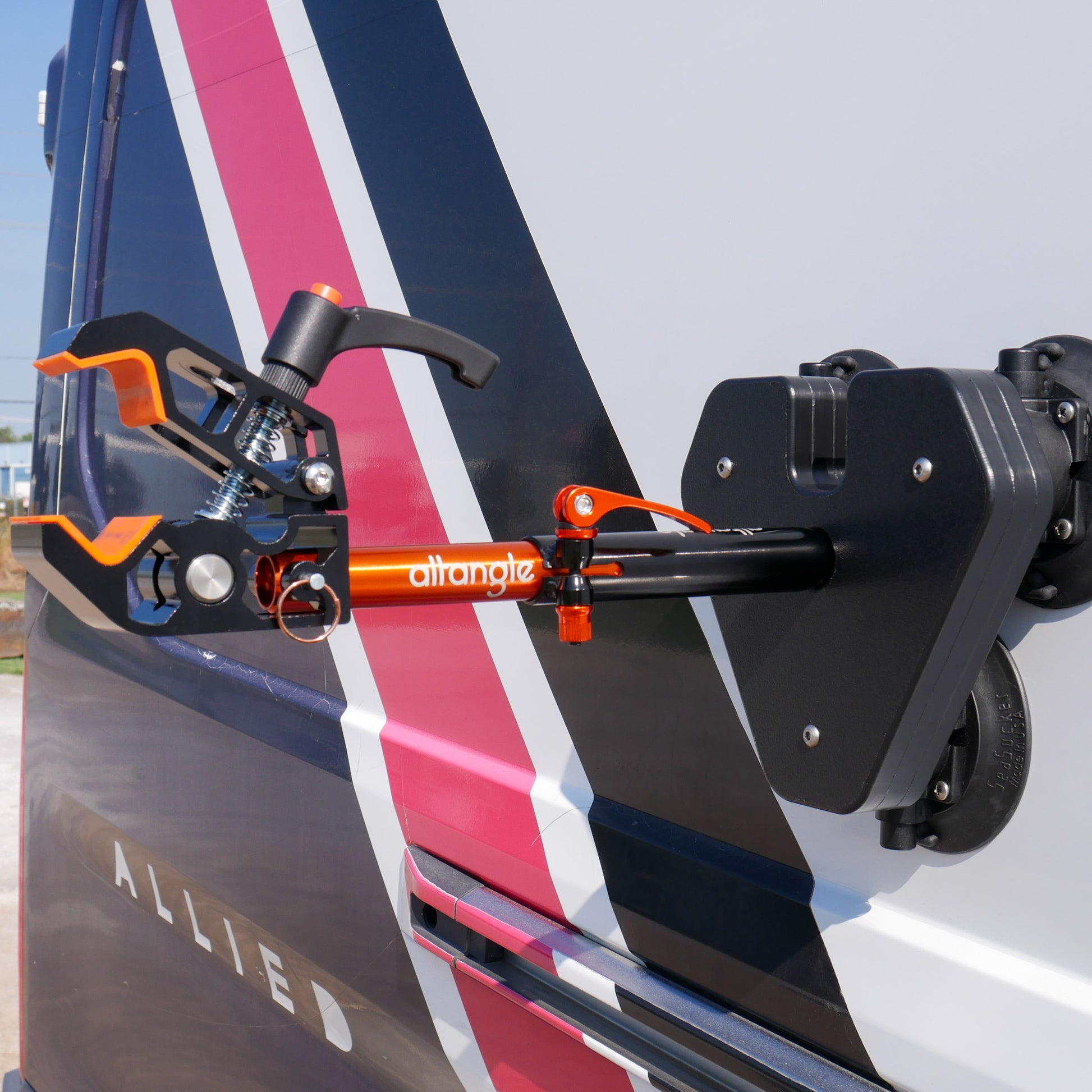 A close-up view of the Connect Air bike stand attached to the side of an Allied vehicle, showcasing its sleek black and orange Altangle clamp mechanism and robust SeaSucker vacuum cup mount. The stand is displayed against the vibrant striped backdrop of the vehicle, emphasizing the product’s detailed engineering and strong visual impact